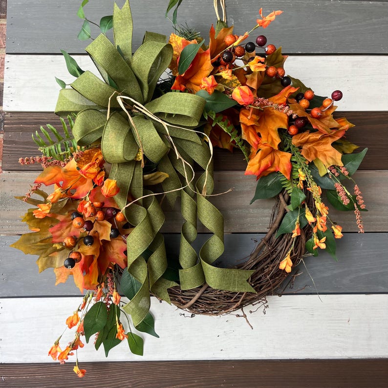 May include: A fall-themed wreath featuring a grapevine base adorned with orange and yellow autumn leaves, berries, and greenery. A large olive green ribbon bow adds a decorative touch to the top of the wreath. The wreath is displayed against a wooden background.