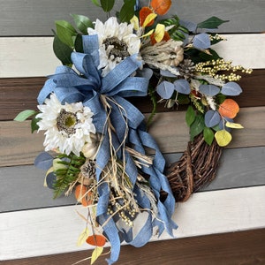 May include: A grapevine wreath decorated with white sunflowers, faux pumpkins, and fall foliage. A large blue ribbon bow is centered on the wreath. The wreath includes green, orange, and yellow leaves, and small white berries.