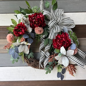 May include: A decorative wreath featuring burgundy peonies, pink pom-poms, and gray-blue eucalyptus leaves. A large black and white striped bow accents the wreath, which is set against a wooden background. The wreath is made of natural grapevine.