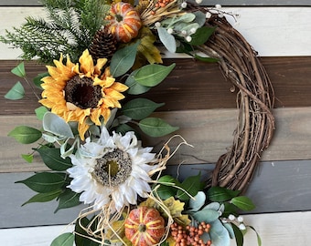 May include: A fall-themed wreath with a grapevine base, adorned with sunflowers, pumpkins, and greenery. The wreath features a mix of yellow, orange, and green elements, creating a rustic and seasonal aesthetic. The wreath is approximately 16 inches in diameter.