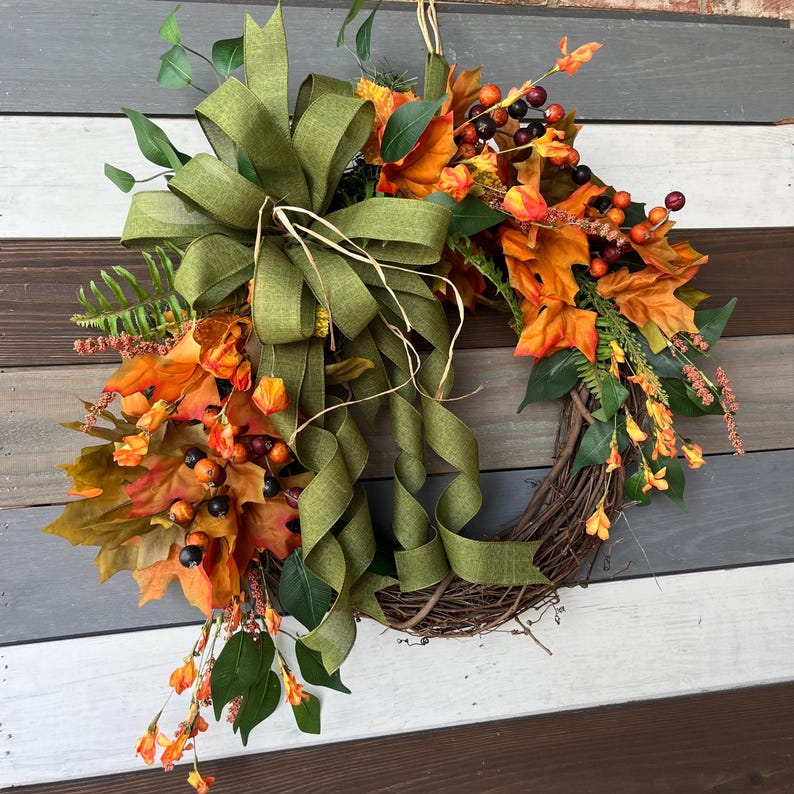 May include: A fall-themed wreath with a grapevine base. It features orange and yellow leaves, black berries, and green foliage. A large olive green bow with long streamers is at the top. The wreath is displayed against a multi-toned wooden background.