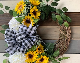 Può includere: Una ghirlanda decorativa con girasoli gialli, fiori bianchi e foglie verdi. Un grande fiocco a scacchi bianco e nero aggiunge contrasto. La ghirlanda è fatta di ramoscelli ed è adatta per la decorazione di porte o pareti.