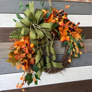 May include: A fall-themed wreath with a grapevine base, adorned with orange and yellow leaves, berries, and greenery. A large olive green bow with long, flowing ribbons adds a decorative touch. Perfect for seasonal home decor.