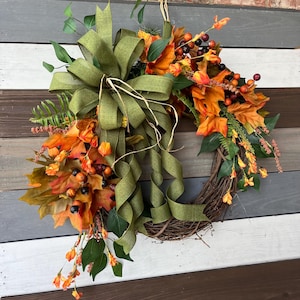 May include: A fall-themed wreath with a woven grapevine base. It features orange and yellow leaves, berries, and greenery, accented by a large olive green bow with long, flowing ribbons. The wreath is designed for seasonal home decor.