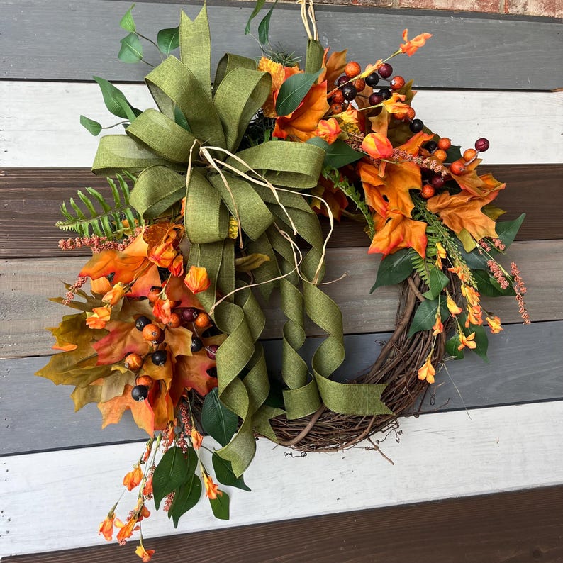 May include: A fall-themed wreath with a woven grapevine base. It features orange and yellow leaves, berries, and greenery, accented by a large olive green bow with long, flowing ribbons. The wreath is designed for seasonal home decor.