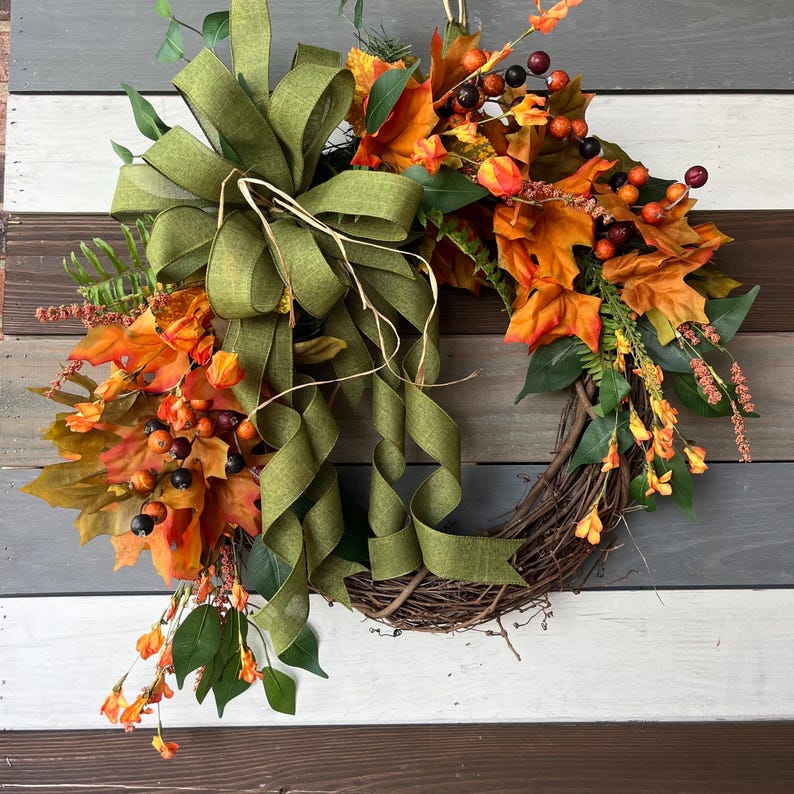 May include: A fall-themed wreath with a grapevine base, adorned with orange and yellow leaves, berries, and sprigs. A large green ribbon bow adds a decorative touch. The wreath is designed for seasonal home decor.
