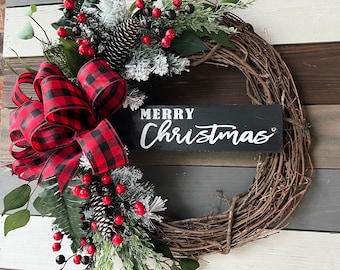 May include: A Christmas wreath with a grapevine base, adorned with red and black plaid ribbon, faux berries, pinecones, and evergreen sprigs. A black sign with the words "MERRY CHRISTMAS" in white is attached to the wreath.