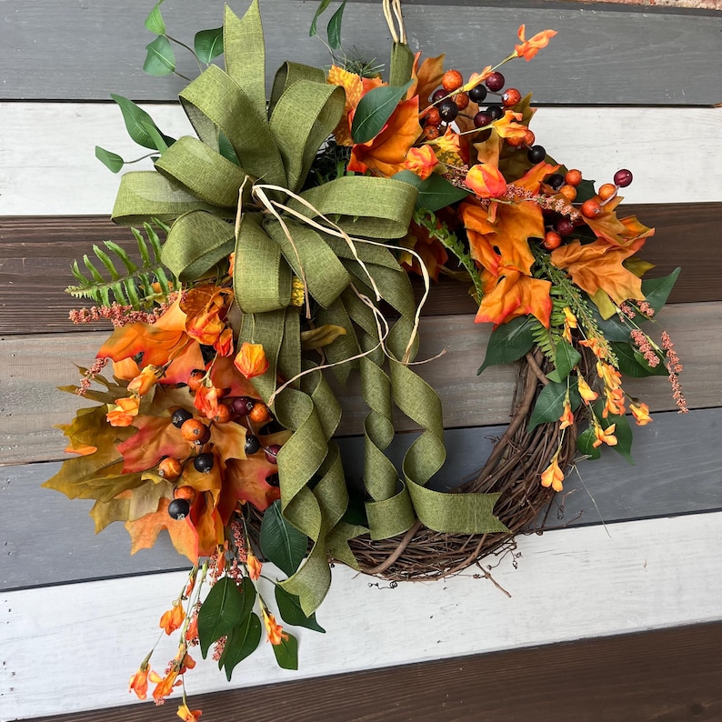 May include: A fall-themed wreath with a woven grapevine base. It features vibrant orange and yellow leaves, berries, and greenery. A large olive green bow with long, flowing ribbons adds a decorative touch to this seasonal home decor item.