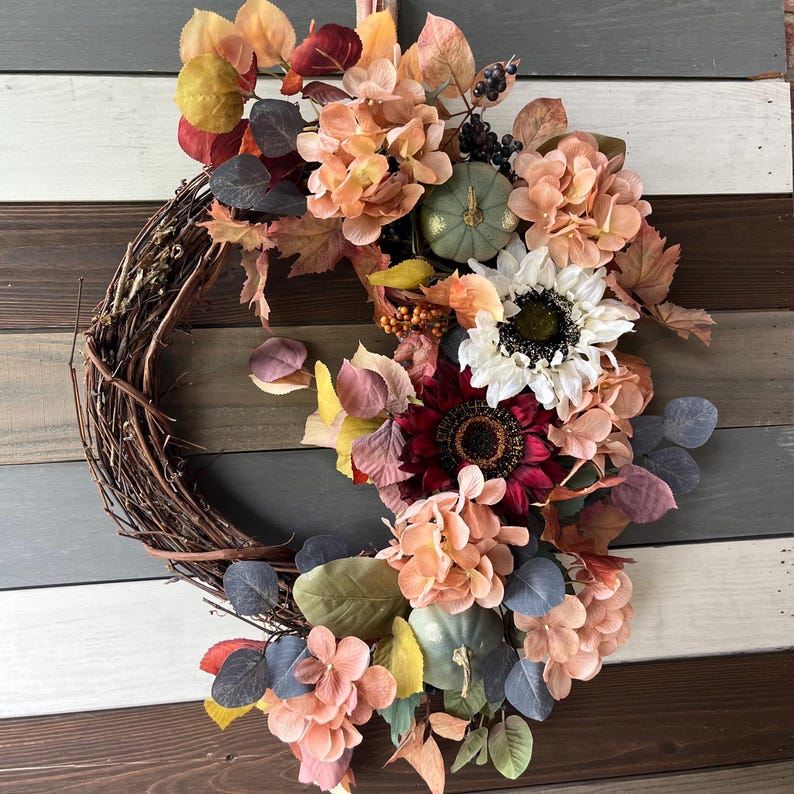 May include: A brown grapevine wreath decorated with peach, burgundy, and white artificial flowers, green leaves, and two small green pumpkins.