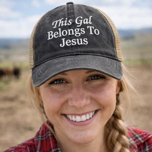May include: A black and tan trucker hat with the text "This Gal Belongs To Jesus" embroidered in white. The hat is worn by a person with blonde hair and a red plaid shirt. The background is a blurred outdoor scene.