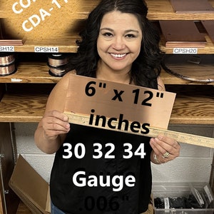 May include: A woman holds a 6 x 12 inch sheet of copper, labeled "COPPER CDA-110 30 32 34 Gauge .006" inches. The copper sheet is on a shelf with other copper products.