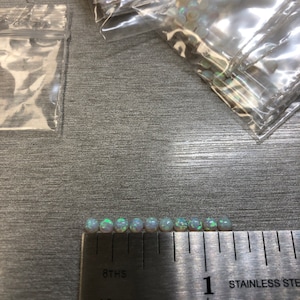 May include: A close-up of a ruler with a row of small, white, iridescent opal beads. The ruler is marked in 1/8ths and 1/16ths of an inch. The beads are approximately 1/4 inch in diameter.