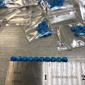 May include: A ruler with markings in 8ths and 16ths of an inch, with the words "Stainless Steel" printed on it. A row of 10 small, blue, iridescent opal beads are lined up on the ruler, measuring approximately 1 inch in length.