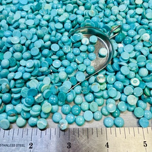 May include: A close-up of a large quantity of turquoise cabochons, a type of gemstone, in various shades of blue and green. The cabochons are spread out on a metal surface with a ruler for scale. A metal scoop is partially filled with cabochons.