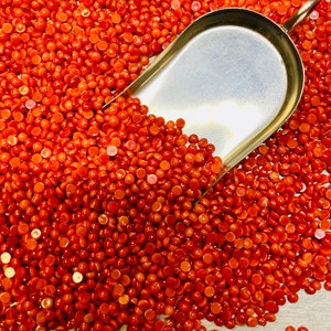 May include: A close-up of a metal scoop filled with small, round, orange beads. The beads are scattered around the scoop, and a ruler is visible at the bottom of the image.
