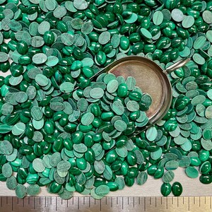 Puede incluir: Una gran cantidad de cabujones ovalados de malaquita verde, algunos de los cuales est&aacute;n en una pala de metal. Los cabujones est&aacute;n dispersos sobre una superficie gris con una regla marcada en pulgadas.