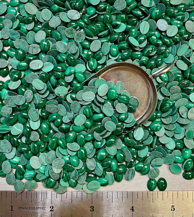 Puede incluir: Una gran cantidad de cabujones ovalados de malaquita verde, algunos de los cuales est&aacute;n en una pala de metal. Los cabujones est&aacute;n dispersos sobre una superficie gris con una regla marcada en pulgadas.