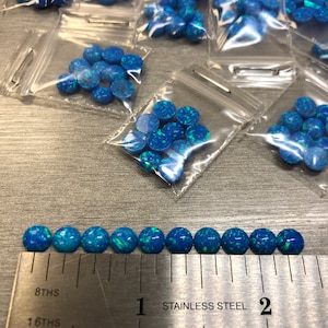 May include: A ruler with markings in inches and 16ths of an inch, with the words "Stainless Steel" printed on it.  A row of 12 small, round, blue opal cabochons are lined up along the ruler, measuring approximately 1 inch in length.