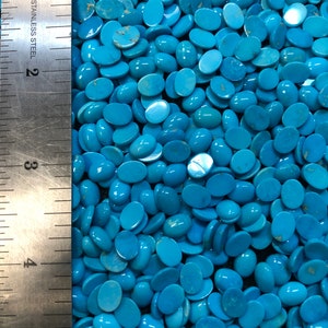 May include: A close-up image of a large quantity of oval-shaped turquoise cabochons. The cabochons are a vibrant blue color and are arranged in a random pattern. A ruler is visible on the left side of the image, indicating the size of the cabochons.