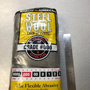 May include: A package of Rhodes American Steel Wool, grade #000, extra fine. The package is yellow with black text and a red star. The steel wool is gray and is packaged in a clear plastic bag. The package also includes a chart showing the different grades of steel wool, from 0000 to 3.