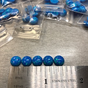 May include: Five blue opal cabochons, each measuring approximately 1/2 inch in diameter, are lined up on a ruler. The ruler is marked in 1/16ths and 1/8ths of an inch. The ruler is labeled "Stainless Steel".
