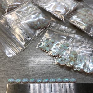 May include: A row of 15 oval-shaped, iridescent white opals, measuring approximately 1 inch in length, are lined up on a ruler. The ruler is marked with inches and the words "Stainless Steel".