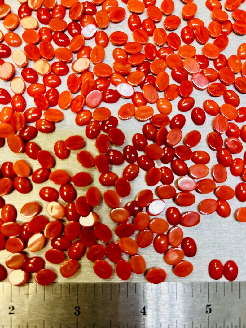 May include: A close-up of a group of oval-shaped, red, and orange gemstone cabochons scattered on a silver surface. A ruler is visible at the bottom of the image, with markings from 2 to 5.