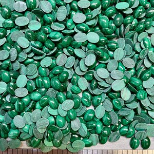 Puede incluir: Una imagen de primer plano de una gran cantidad de cabujones ovalados de malaquita verde. Los cabujones est&aacute;n dispersos sobre una superficie met&aacute;lica con una regla para la escala. La regla muestra medidas en pulgadas.