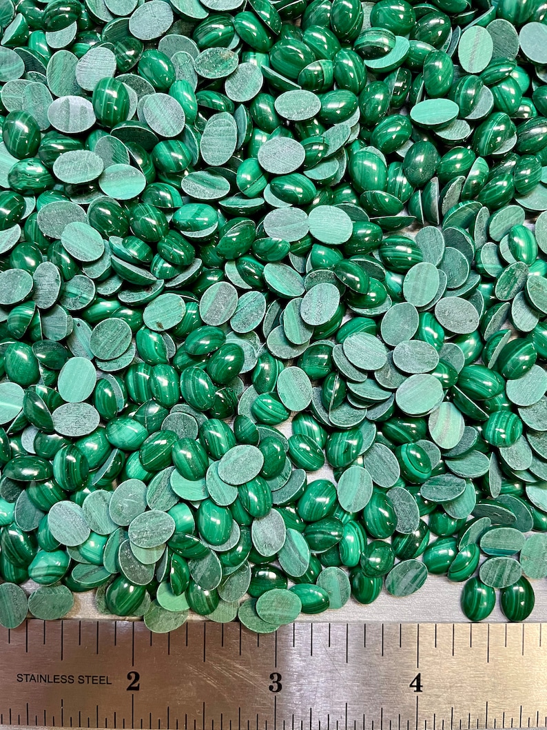 Puede incluir: Una imagen de primer plano de una gran cantidad de cabujones ovalados de malaquita verde. Los cabujones est&aacute;n dispersos sobre una superficie met&aacute;lica con una regla para la escala. La regla muestra medidas en pulgadas.