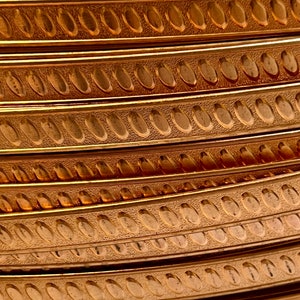 May include: Close-up of a stack of copper-colored metal trim with a repeating oval pattern. The trim has a textured surface.