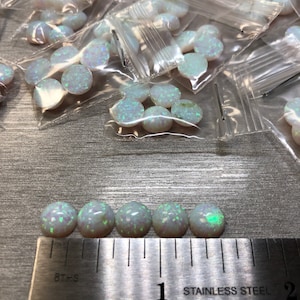 May include: A close-up of six white opal cabochons with green and blue iridescence, lined up on a ruler measuring 1 inch. The ruler is made of stainless steel and has markings for 8ths and 16ths of an inch.