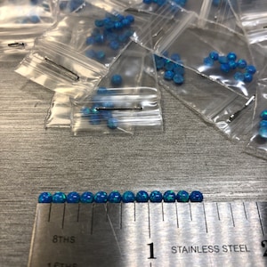 May include: A ruler with a stainless steel measuring tape marked in 8ths and 16ths of an inch. A row of 20 small, round, blue opal gemstones are lined up along the ruler, measuring approximately 1 inch in length.