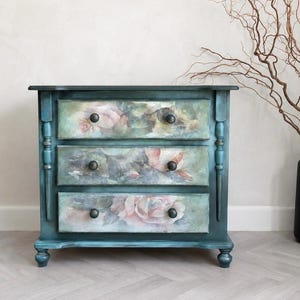 May include: A three-drawer chest finished in a deep teal blue with a distressed texture. Each drawer front features a soft, muted floral print of pink and white roses. The piece includes dark, round knobs and decorative turned side columns.