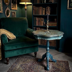 May include: A dark green velvet armchair with a gold patterned throw blanket, positioned next to a small, round, blue-painted pedestal table with a scalloped edge and a brass candlestick holder, set against a dark teal wall with a bookshelf.