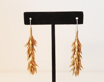 May include: A pair of gold-colored dangle earrings, each featuring a textured, elongated design resembling a wheat stalk. The earrings are displayed on a black velvet jewelry stand against a white background. The earrings are suspended from silver-colored hooks.
