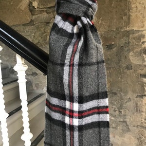 Peut inclure: Une écharpe à carreaux gris, noir et rouge avec des franges. L'écharpe est pliée et drapée sur une rampe. L'étiquette sur l'écharpe indique "House of Tartan".