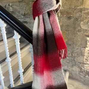May include: A long rectangular scarf featuring a plaid pattern in shades of red, cream, brown, and grey. The scarf has a soft, woven texture with fringed ends and a small rectangular white label stitched onto the bottom corner of the fabric.