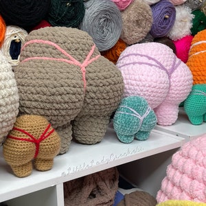 May include: A collection of crocheted butt pillows in various colors, including brown, pink, teal, and tan. The pillows are arranged on a white shelf with a sign that reads "@littleladycrochet".