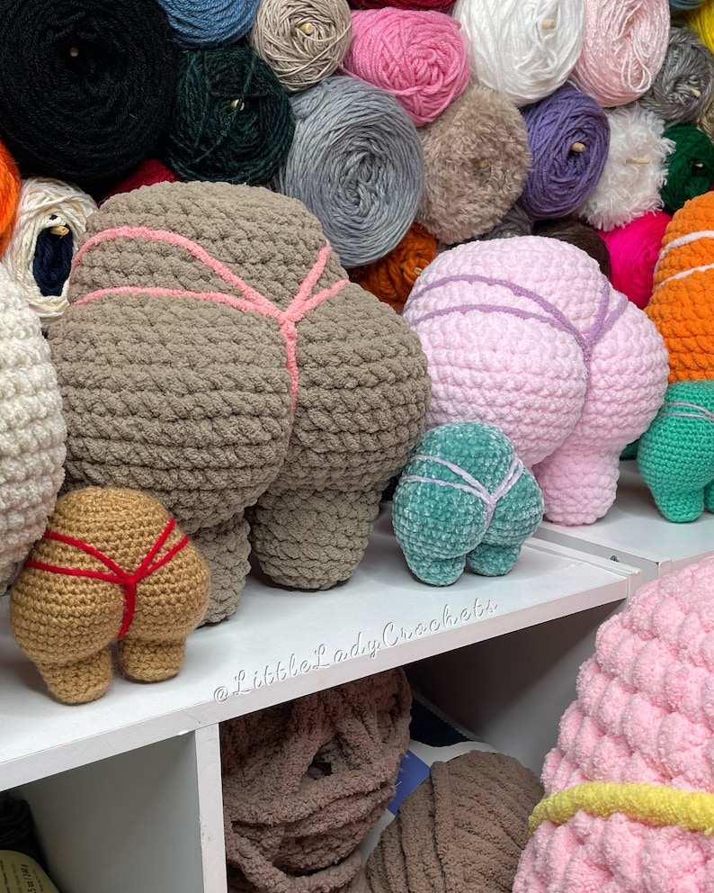 May include: A collection of crocheted butt pillows in various colors, including brown, pink, teal, and tan. The pillows are arranged on a white shelf with a sign that reads "@littleladycrochet".