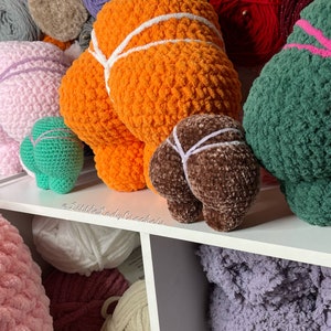 May include: A collection of crocheted plush toys in various colors, including orange, brown, green, and pink. The toys are arranged on a white shelf with balls of yarn in the background. The text "@LittleLadyCrockers" is visible on the shelf.