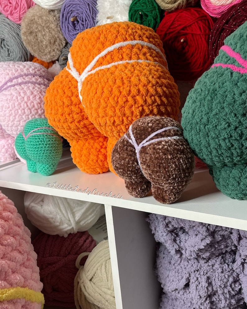 May include: A collection of crocheted plush toys in various colors, including orange, brown, green, and pink. The toys are arranged on a white shelf with balls of yarn in the background. The text "@LittleLadyCrockers" is visible on the shelf.
