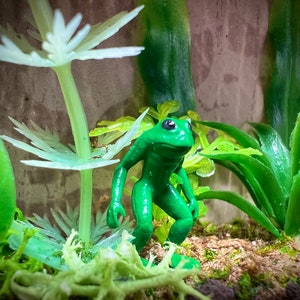 May include: A vibrant green frog figurine stands amidst artificial plants and moss. The frog has black eyes and is posed as if it is standing. The background features more greenery and a textured, neutral-toned surface.