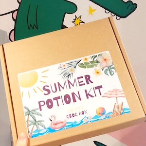 May include: A brown cardboard box with a label that reads "Summer Potion Kit." The label features a sun, palm trees, a flamingo float, and a beach scene with an umbrella and beach chair. The words "CRISPY CROC BOX" are also on the label.