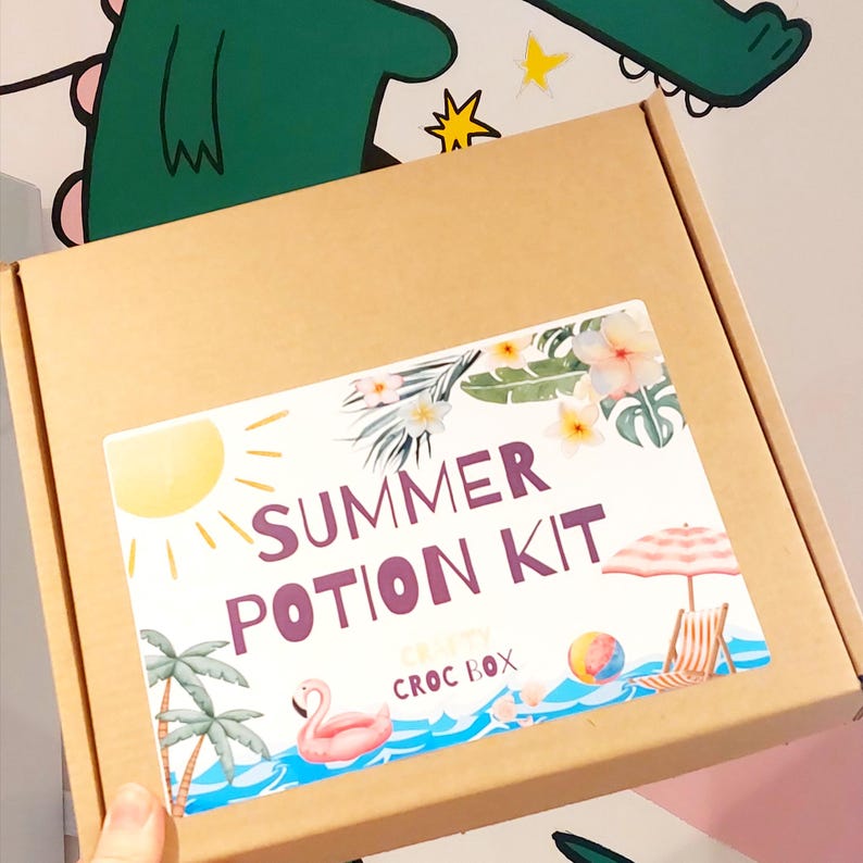 May include: A brown cardboard box with a label that reads "Summer Potion Kit." The label features a sun, palm trees, a flamingo float, and a beach scene with an umbrella and beach chair. The words "CRISPY CROC BOX" are also on the label.