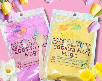 May include: Two clear pouches of bath fizz, one pink and one yellow, with wooden spoon. The pink pouch reads "EGGstra Magic" and the yellow pouch reads "EGGstra Fizzy Magic". The background features flowers.