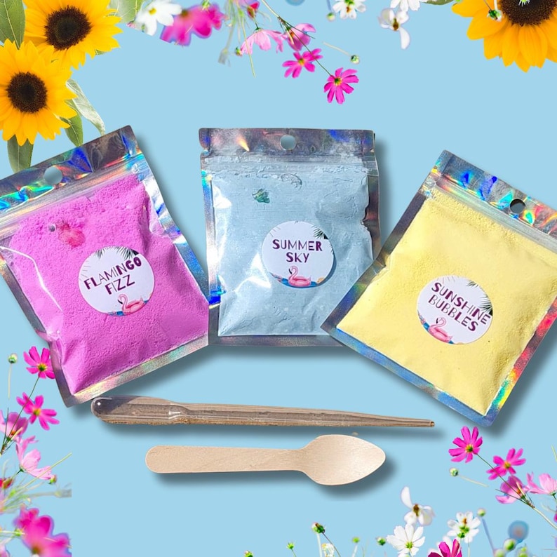 May include: Three colorful bath fizz packets labeled "Flamingo Fizz", "Summer Sky", and "Sunshine Bubbles" with a wooden spoon. The packets are pink, blue, and yellow, respectively, and are in clear, shiny pouches. The background is light blue with flowers.