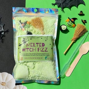 May include: A bag of green bath fizz labelled "Melted Witch Fizz" with witch-themed accessories. The image features miniature witch hats, brooms, and a green witch face. The background is green and black, with a white pumpkin.