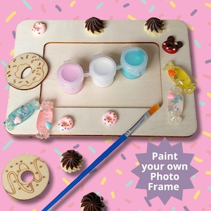 May include: A wooden photo frame with a paint-by-numbers design. The frame is decorated with donuts, candy, and chocolate swirls. The kit includes three small bottles of paint and a paintbrush. The text "Paint your own Photo Frame" is printed on the image.