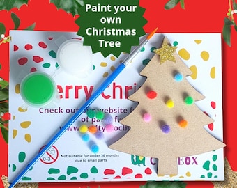 Op de afbeelding: Een doe-het-zelf kerstboom knutselset met een houten boomuitsparing, verf, een kwast en kleurrijke pompoenen. De set bevat de tekst "Verf je eigen kerstboom" en "Kijk op onze website voor meer creatieve opties".