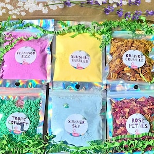 May include: A collection of colorful bath products in clear pouches, including pink "Flamingo Fizz", yellow "Sunshine Bubbles", and orange "Orange Crumble". Other pouches contain green confetti, blue "Summer Sky", and rose petals. Each pouch has a circular label with a flamingo.